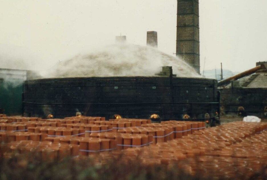 A domed beehive kiln firing bricks, with a tall chimney behind it.